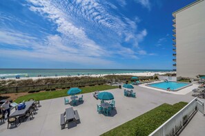 Ein Poolbereich mit Liegestühlen und Sonnenschirmen mit Blick auf einen Strand, Menschen und einen klaren Himmel.