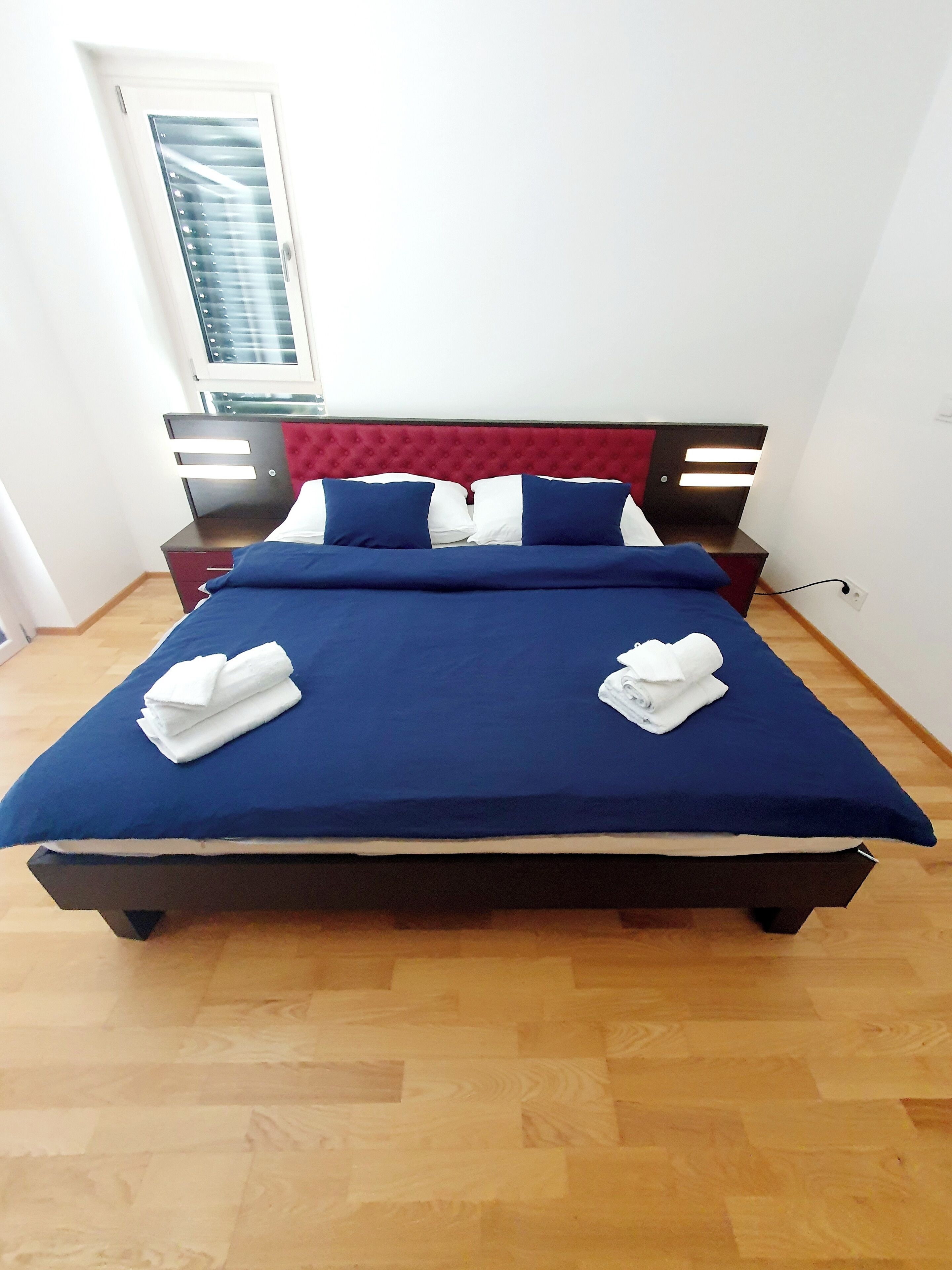 Ein ordentlich bezogenes Bett mit blauer Bettwäsche, weißen Kissen, einem roten Kopfteil und zwei Nachttischen mit Lampen.