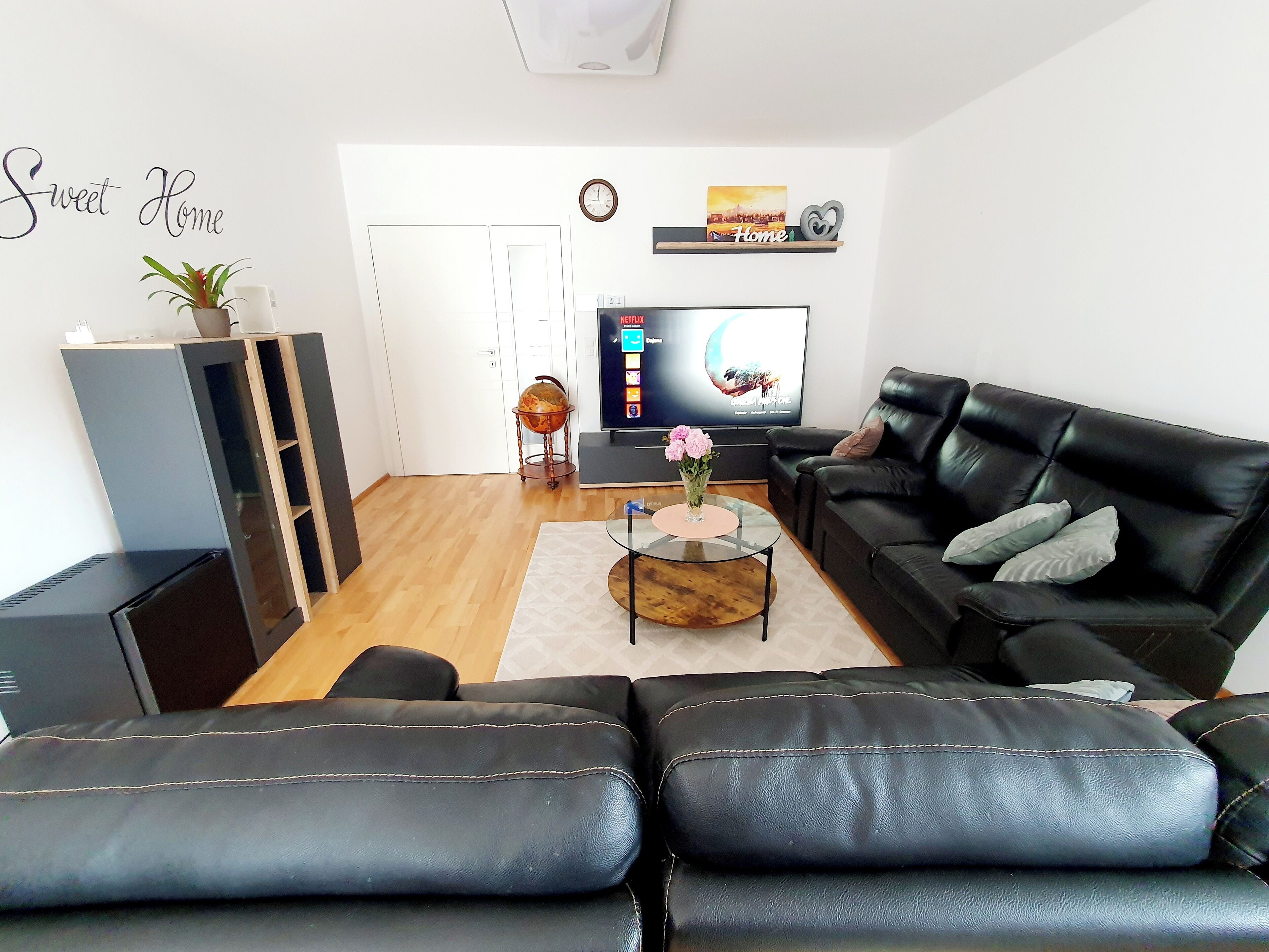 Ett modernt vardagsrum med en svart lädersoffa, ett soffbord i glas och en platt-TV. På ett stativ står en liten jordglob, och det finns även en krukväxt och en hylla med ett fotoram och en hjärtformad prydnad.