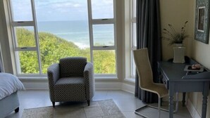 A room with a large window offering a view of the ocean, a patterned armchair, a desk with a chair, and a bed with a blue headboard. - Sunshine Modern Retreat (Port Alfred)