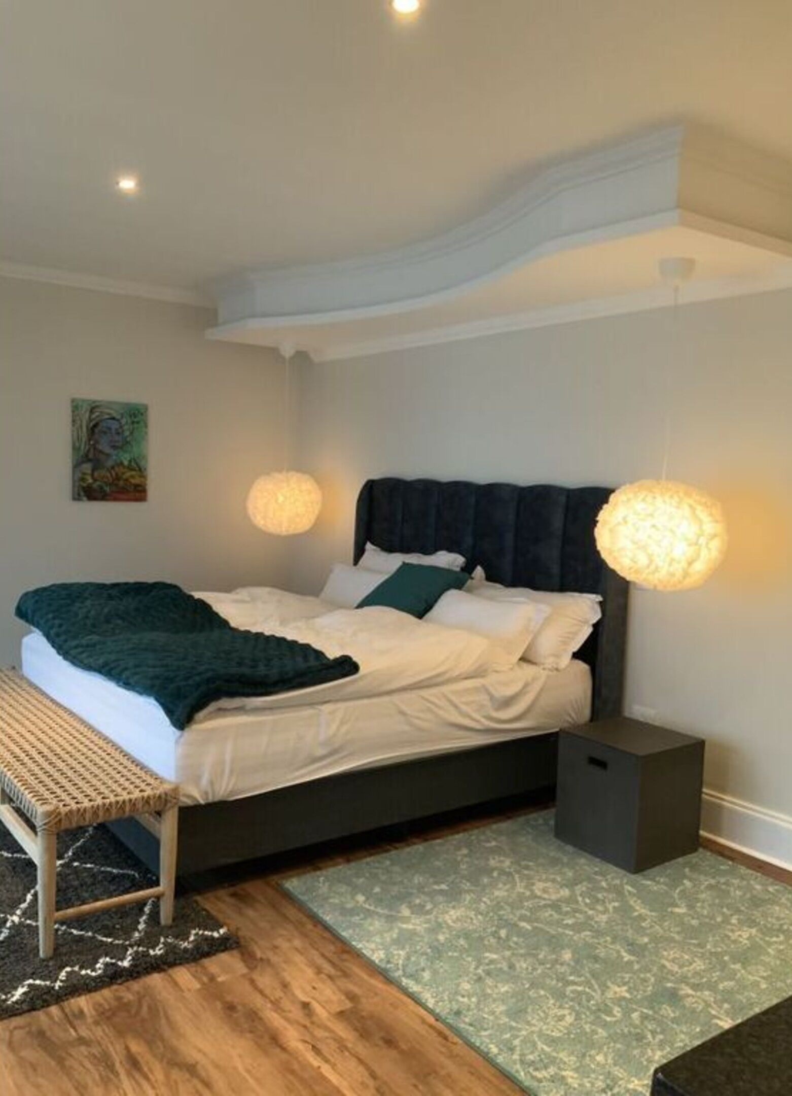 Un dormitorio con cama, mesitas de noche, un banco, una alfombra y un cuadro en la pared.