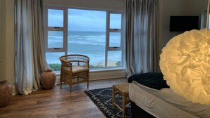 A bedroom with a large window overlooking the sea, a wicker chair, a bed with a white headboard, and a flat-screen TV on the wall. - Sunshine Modern Retreat (Port Alfred)