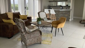 A modern living room with a brown sofa, wicker chairs, a coffee table, and a kitchen area in the background. - Sunshine Modern Retreat (Port Alfred)