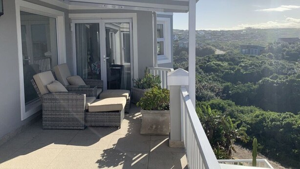 A balcony with a wicker sofa and cushions, a potted plant, and a view of a green landscape. - Sunshine Modern Retreat (Port Alfred)