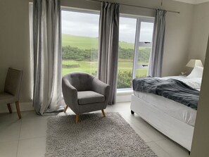 A bedroom with a bed, a chair, and a view of a green landscape through large windows. - Sunshine Modern Retreat (Port Alfred)
