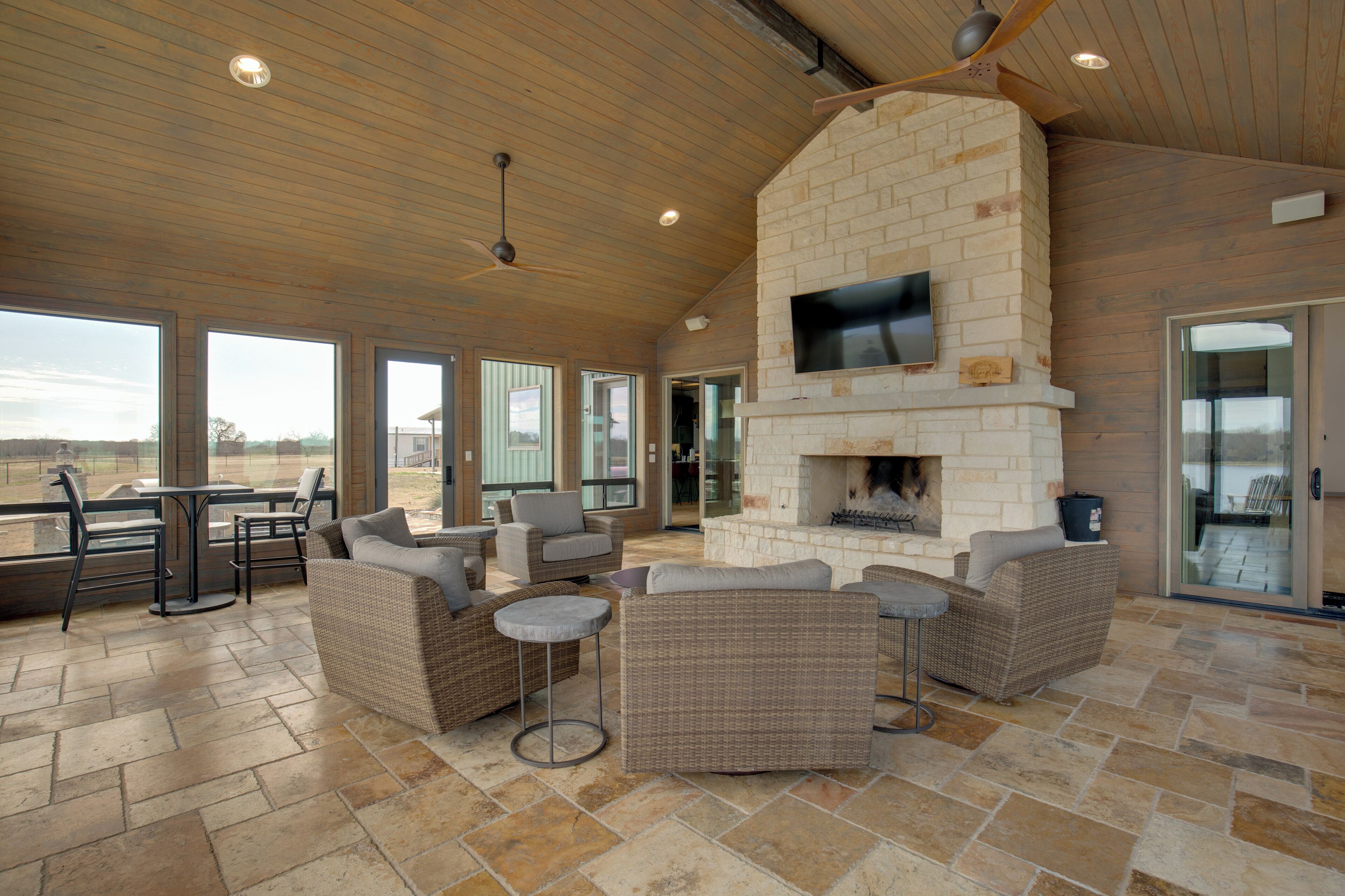 A spacious outdoor lounge area with wicker furniture, a stone-tiled floor, and a stone fireplace.