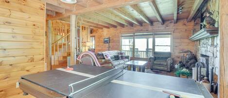 A wooden cabin interior with a ping pong table, a staircase, a fireplace, and a cozy seating area.