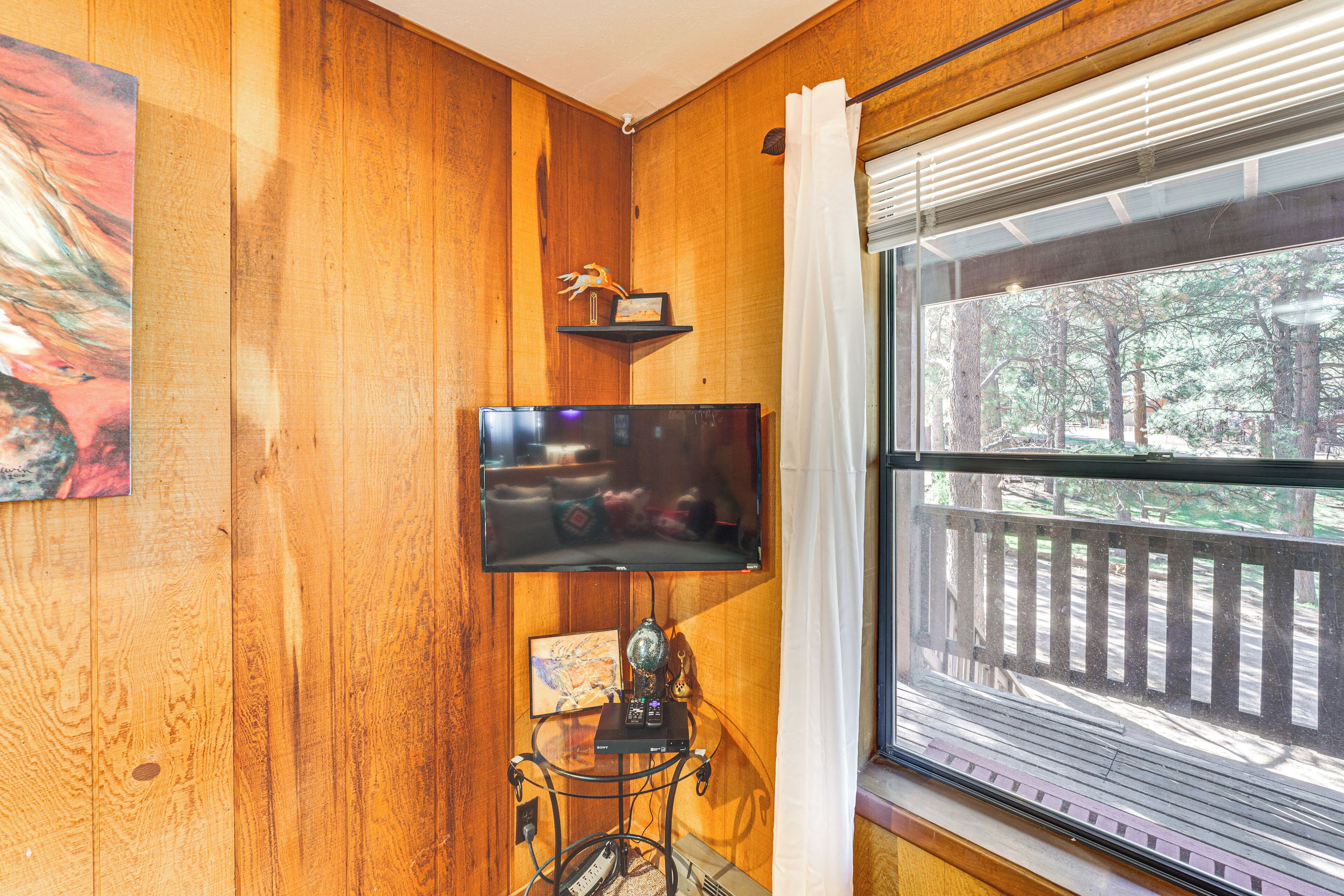 A wooden-paneled room with a TV mounted on the wall, a small shelf with decorative items, and a window with a view of trees.