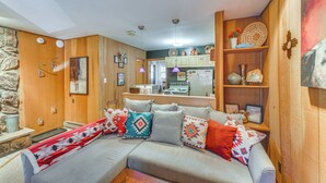 A cozy living room with a sectional sofa, wooden walls, and a kitchen in the background. - Easy Resort Access: Angel Fire Adventure Condo! (Angel Fire)