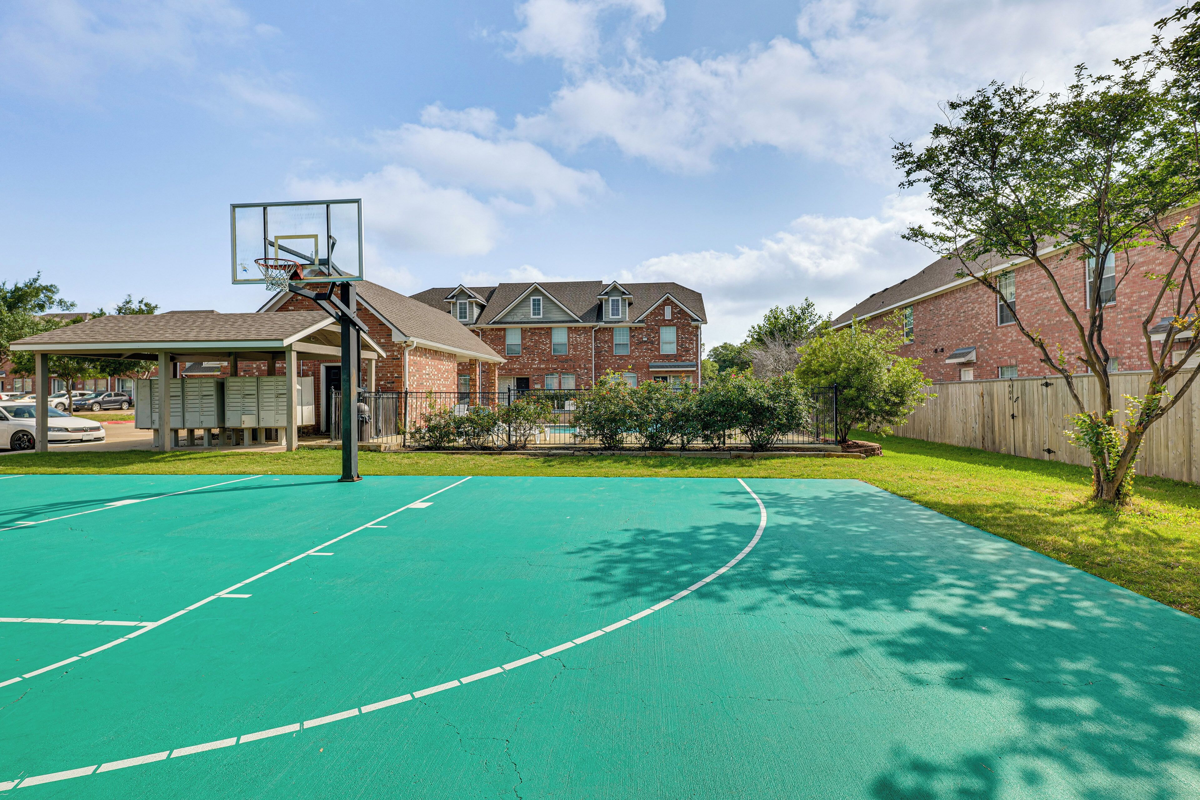Un terrain de basket avec un panier, entouré d’un bâtiment en briques et d’une clôture en bois.