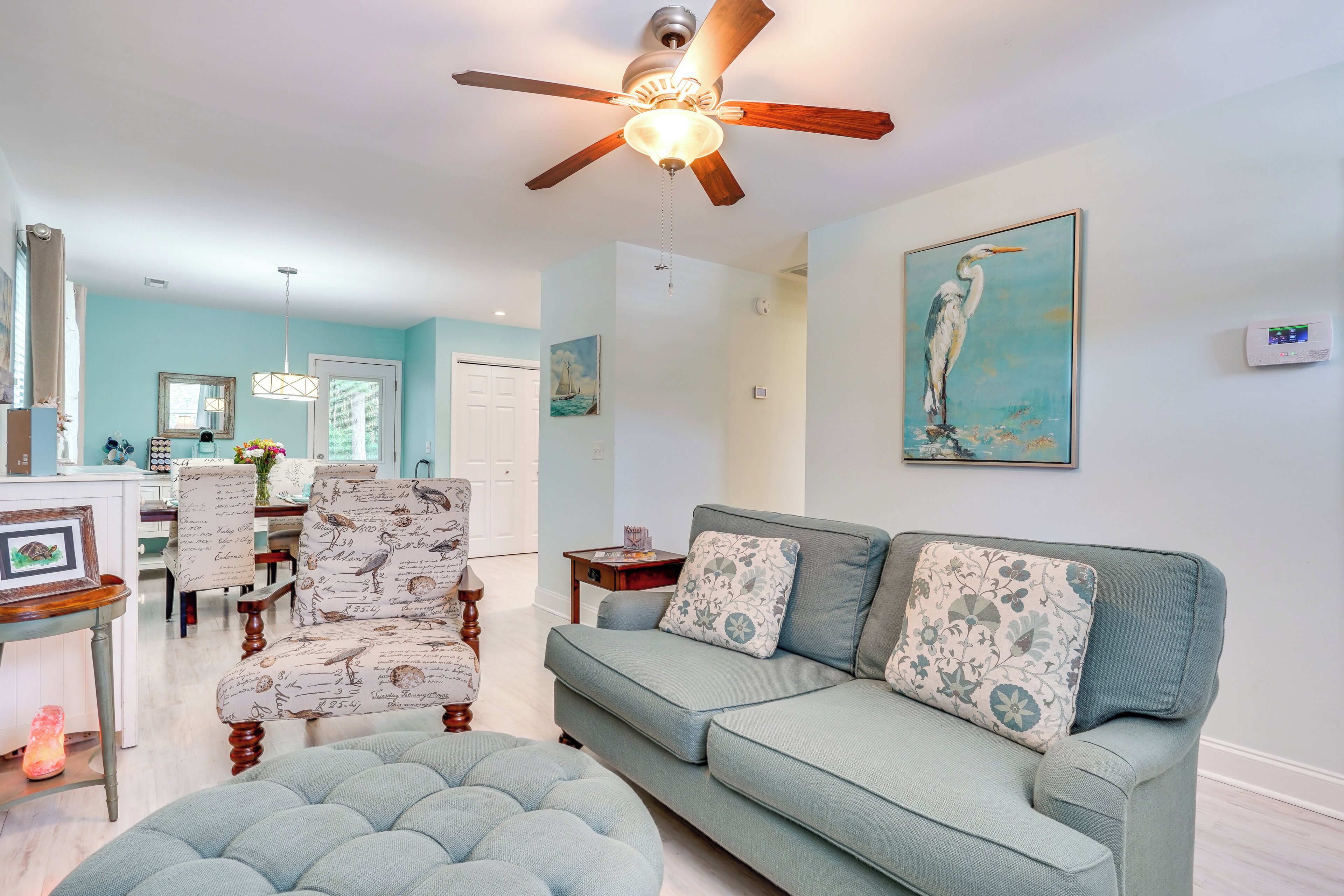 A living room with a light blue sofa, patterned armchairs, a ceiling fan, and a painting of a bird on the wall.