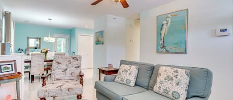 A living room with a light blue sofa, patterned armchairs, a ceiling fan, and a painting of a bird on the wall.