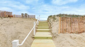 A wooden staircase leading to a beach area with a white railing, surrounded by sand and a wooden fence.