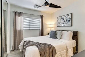 Une chambre à coucher comprenant un lit, un ventilateur de plafond, une fenêtre avec des stores, une œuvre d’art accrochée au mur et une table de chevet avec une lampe.