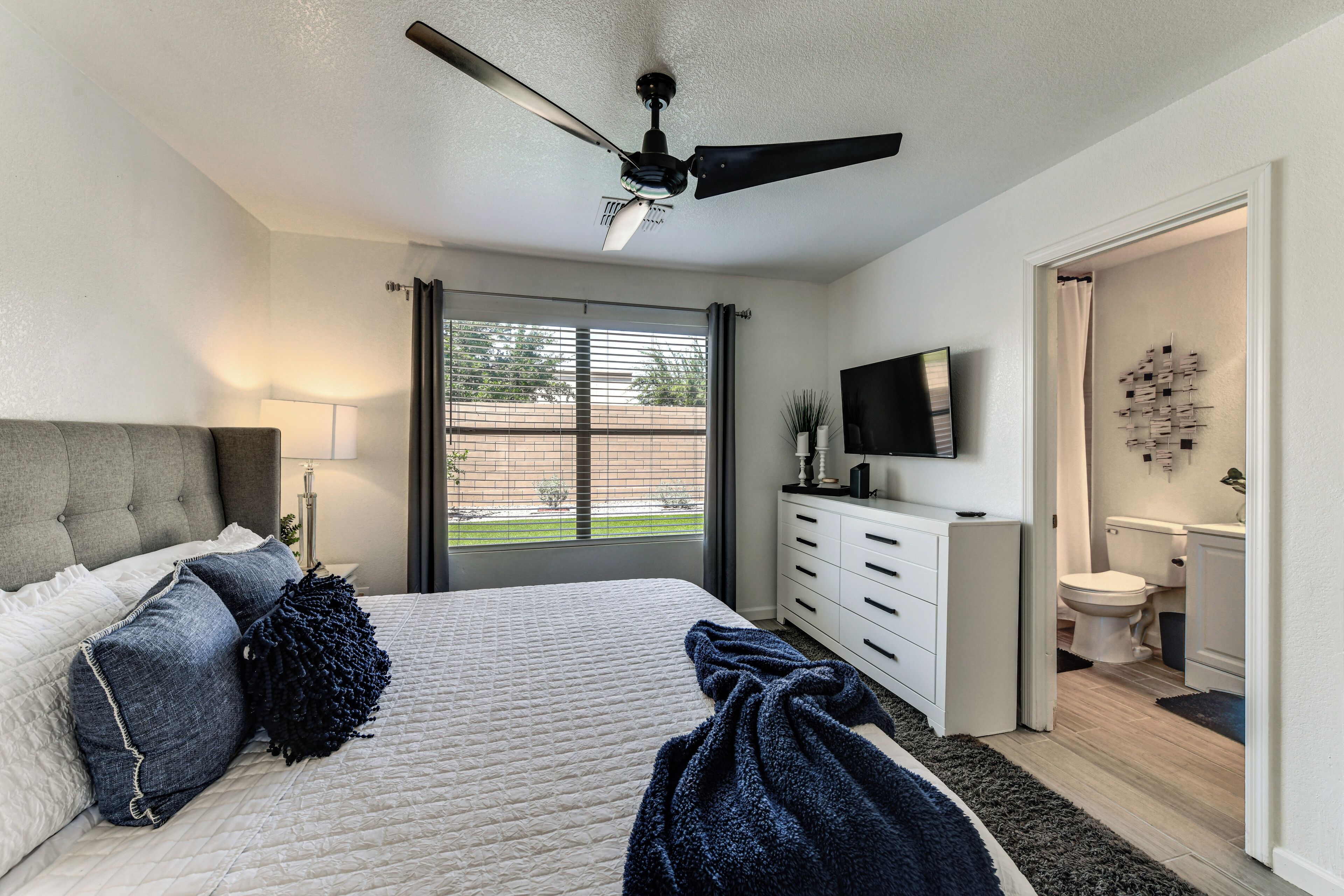 Quarto com cama, ventilador de teto, janela com persianas, cômoda, televisão e banheiro.