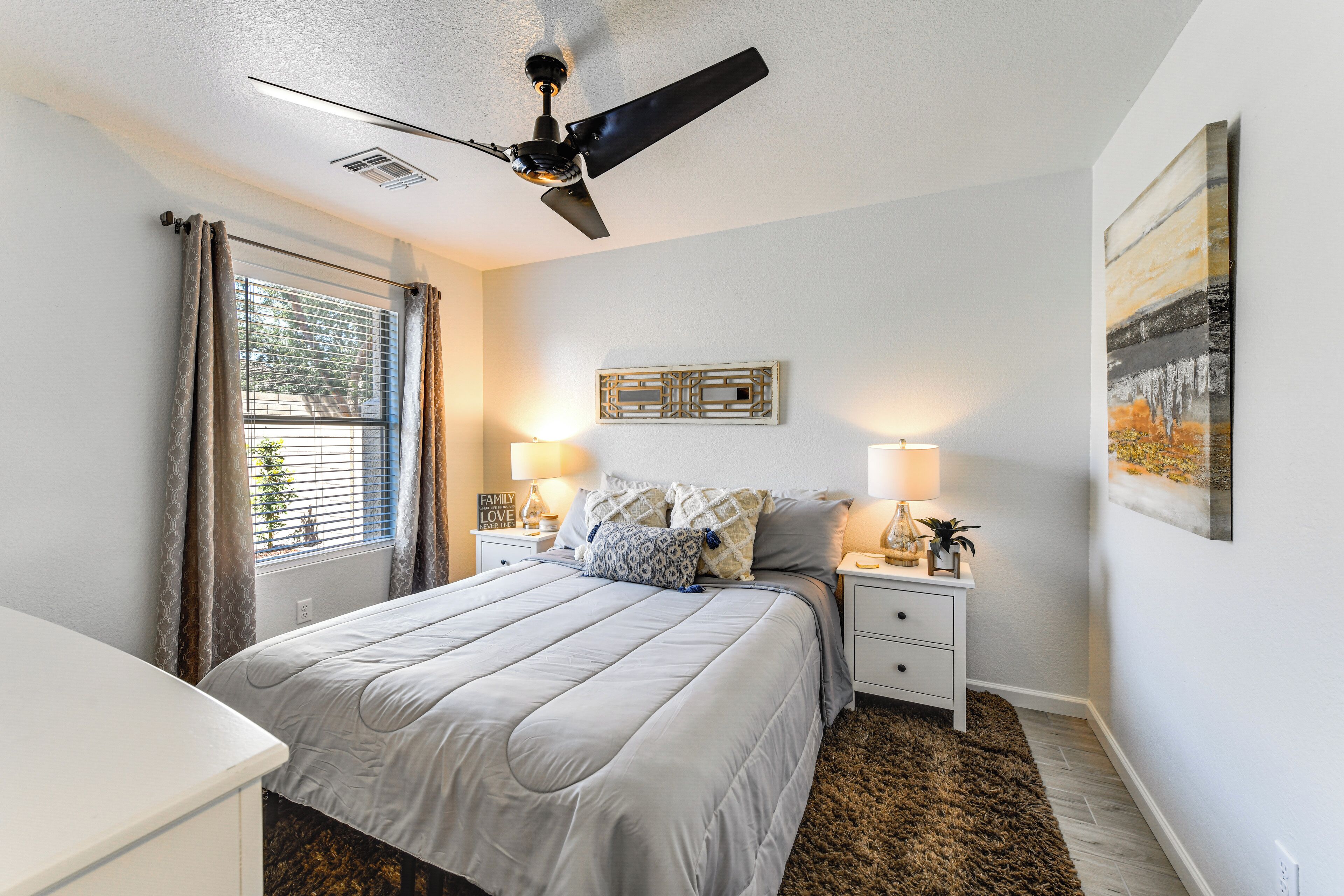 A bedroom with a bed, nightstands, lamps, a ceiling fan, a window with curtains, and a wall-mounted artwork.