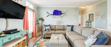 A living room with a sectional sofa, a flat-screen TV mounted on the wall, a ceiling fan, and a colorful rug.