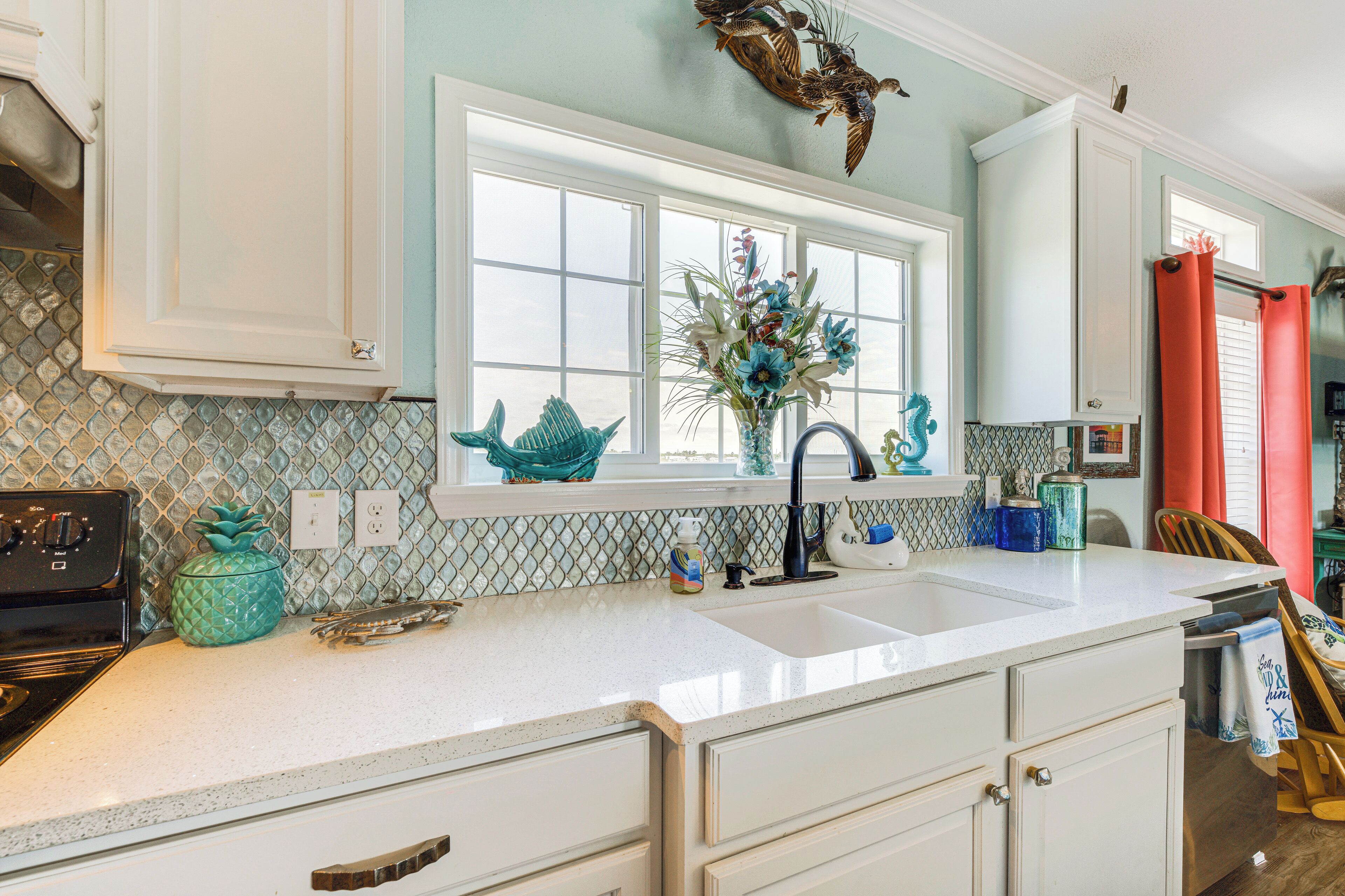 Une cuisine avec des meubles blancs, une grande fenêtre et une sculpture de poisson décorative accrochée au mur.