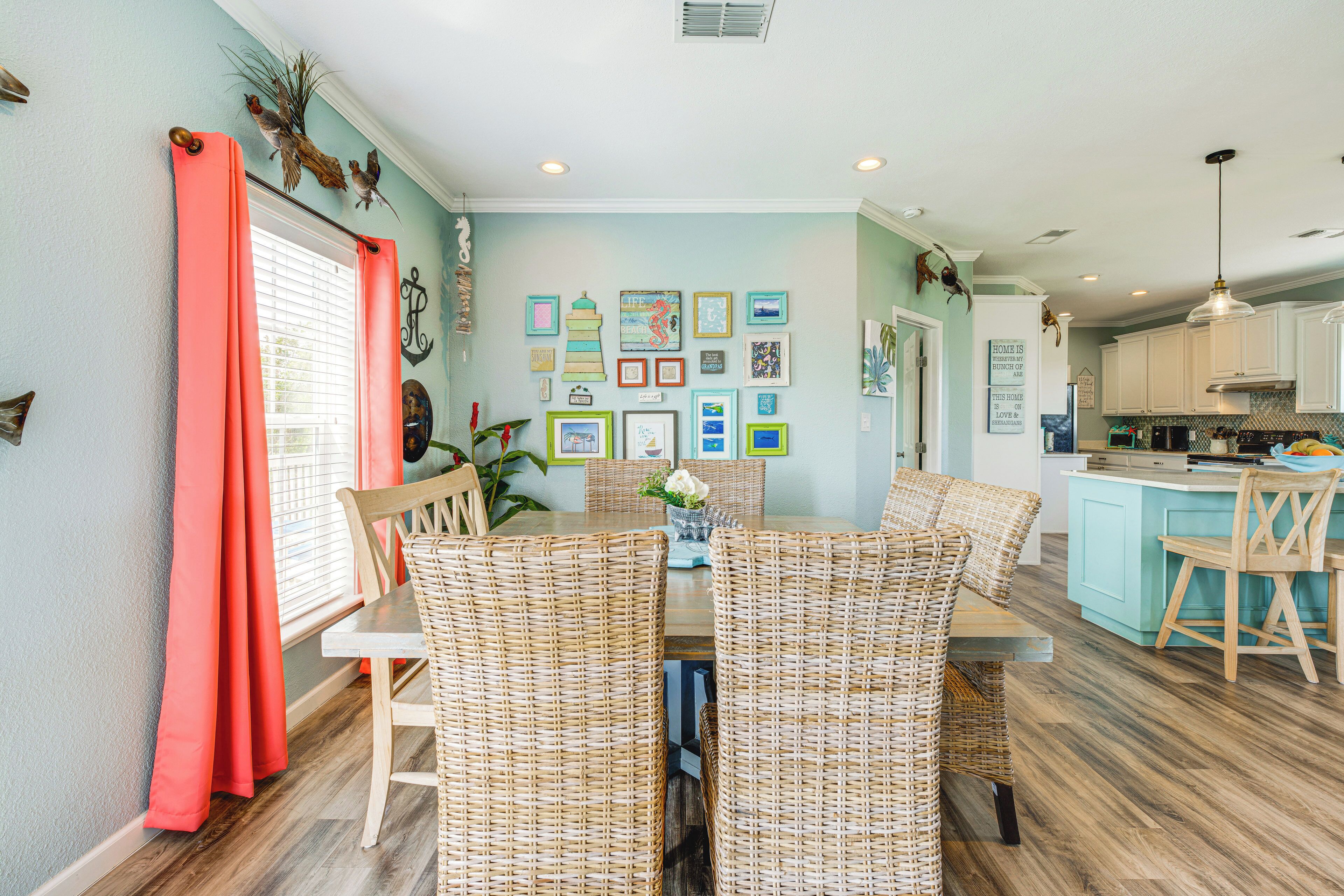 A dining area with wicker chairs and a table, a kitchen with white cabinets, and a wall decorated with framed pictures.