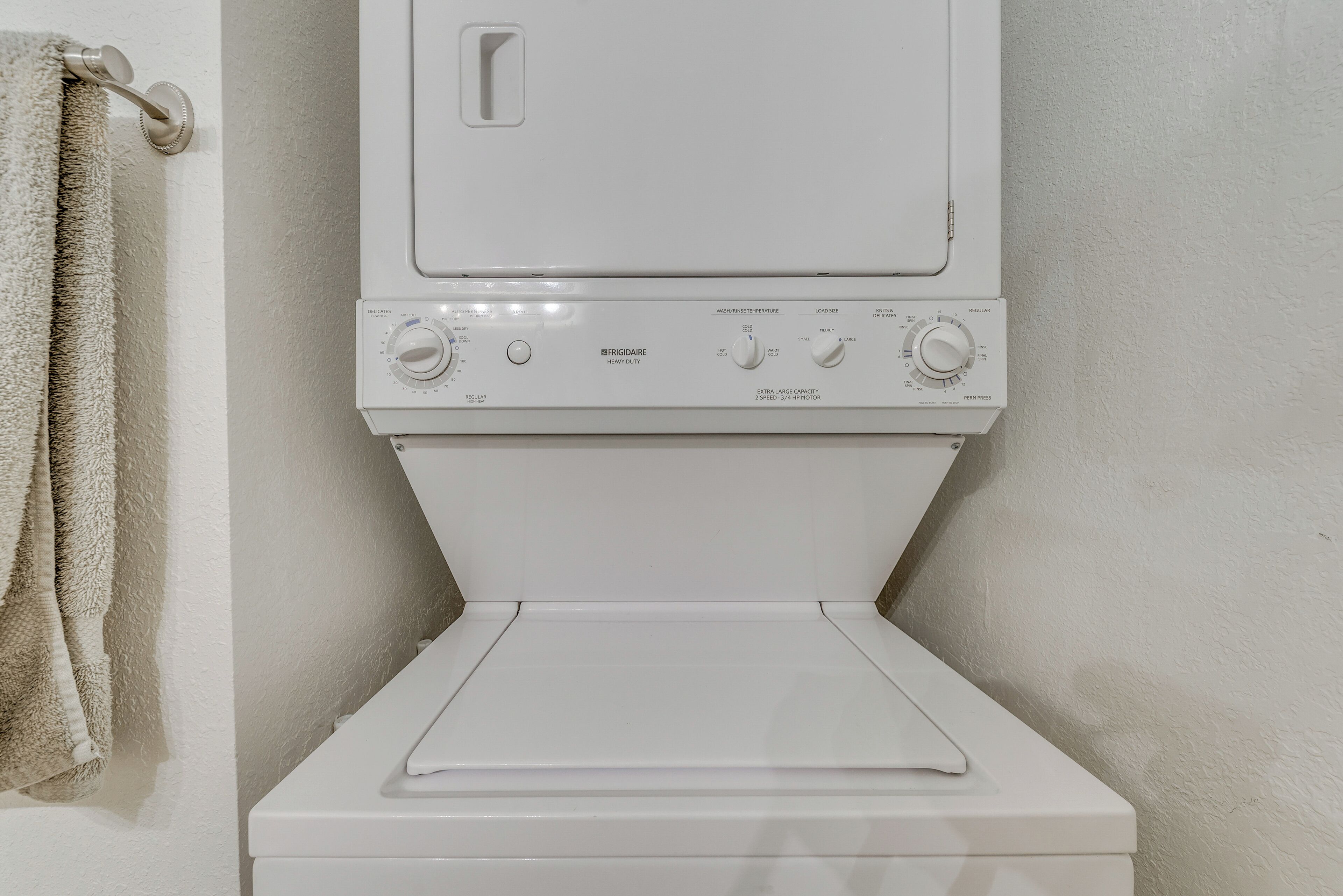 A front view of a white washing machine with control knobs and a front-loading door, installed in a bathroom with a towel hanging on a rack.