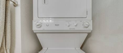 A front view of a white washing machine with control knobs and a front-loading door, installed in a bathroom with a towel hanging on a rack.