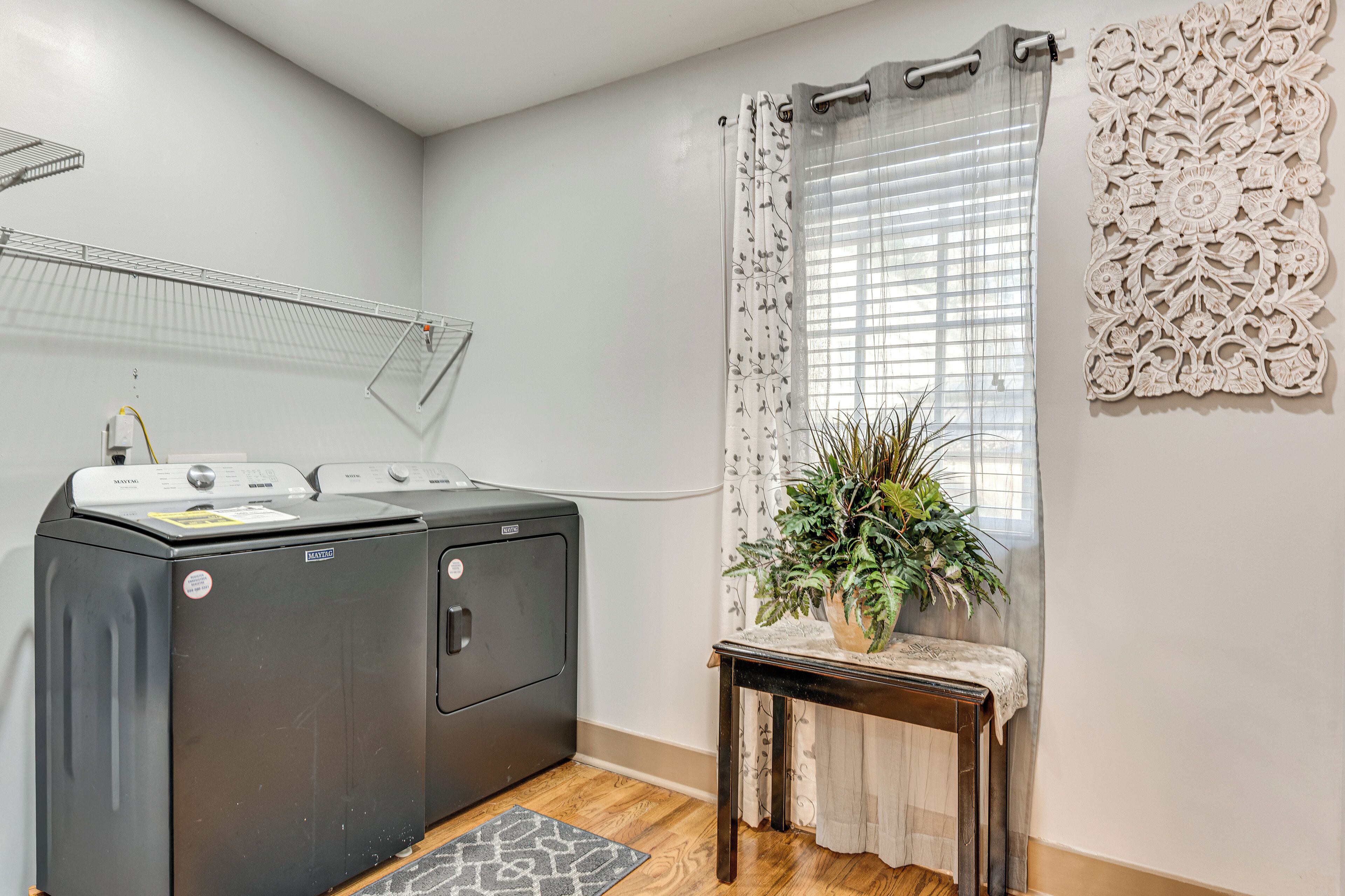 Un área de lavandería con lavadora y secadora de carga frontal, una pequeña mesa con una planta y una ventana con cortinas decorativas.