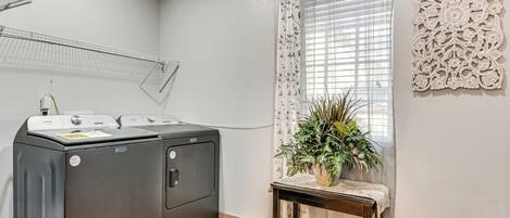 Un área de lavandería con lavadora y secadora de carga frontal, una pequeña mesa con una planta y una ventana con cortinas decorativas.