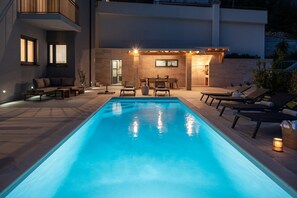 Une piscine dotée de chaises longues et d’une table, entourée d’un patio moderne et agréé d’une entrée bien éclairée.