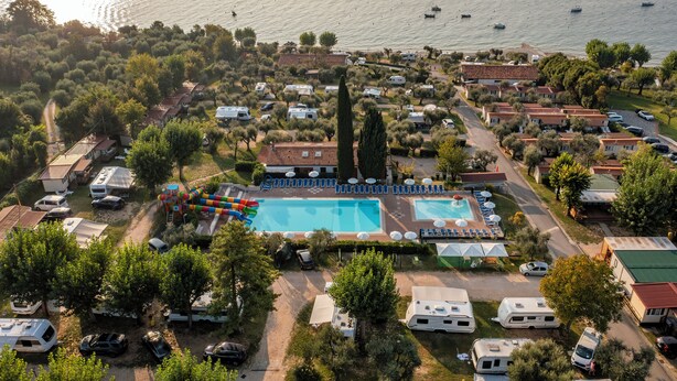 An aerial view of a vacation area with a swimming pool, RVs, and boats on the water. - Camping Fontanelle (Moniga del Garda)