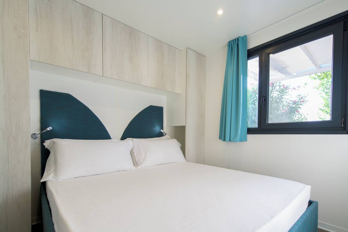 Una cama bien hecha con cabecera, una ventana con cortinas y vista a follaje exterior.