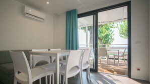 A modern dining area with a white table and chairs, a green sofa, and a sliding glass door leading to an outdoor patio with a view of trees. - Camping Fontanelle (Moniga del Garda)