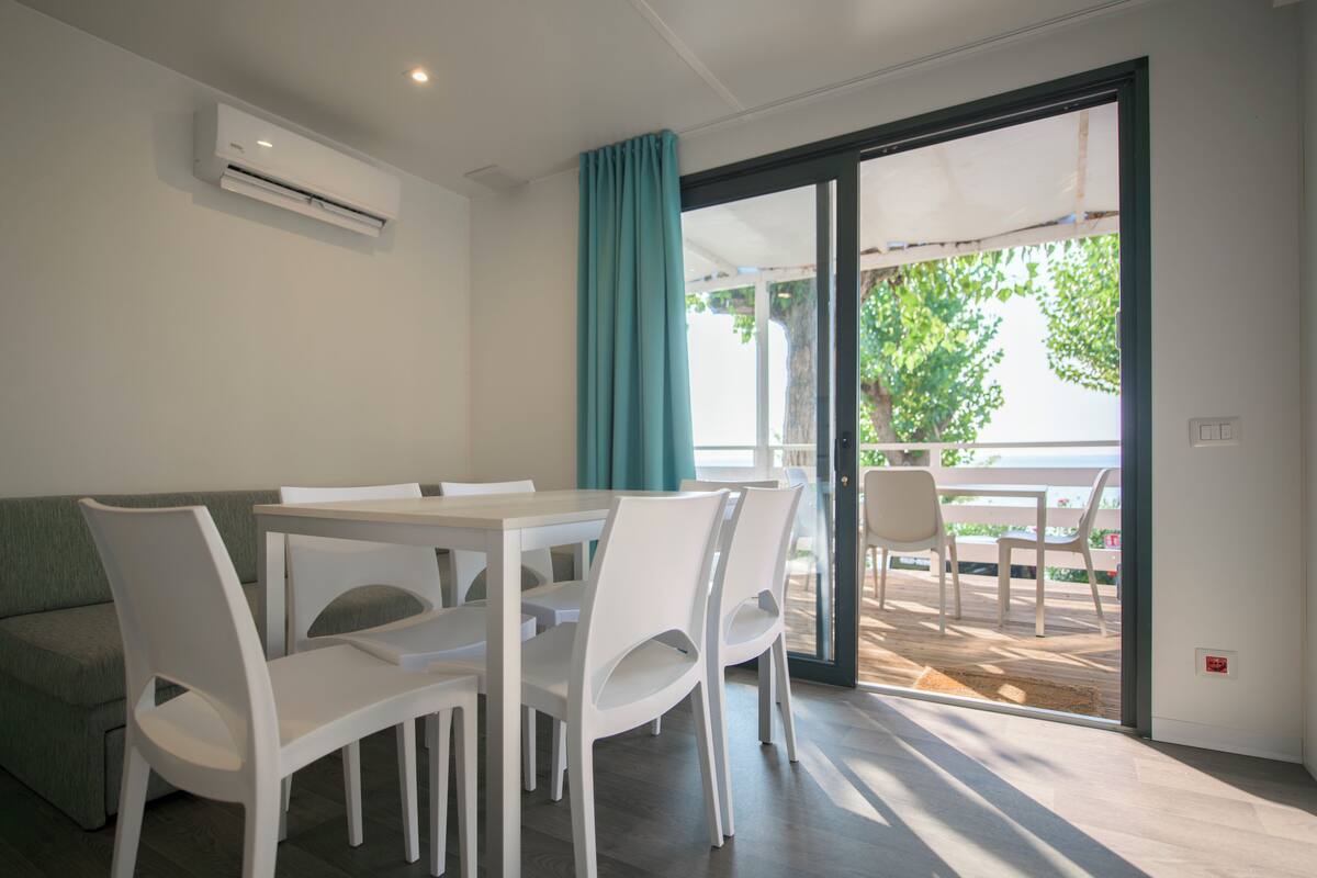 A modern dining area with a white table and chairs, a green sofa, and a sliding glass door leading to an outdoor patio with a view of trees.
