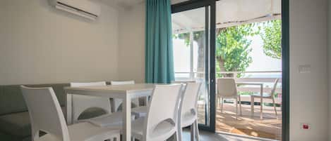 Un comedor moderno con una mesa y sillas blancas, un sofá verde y un ventanal corredero que da a un patio exterior con vistas a árboles.