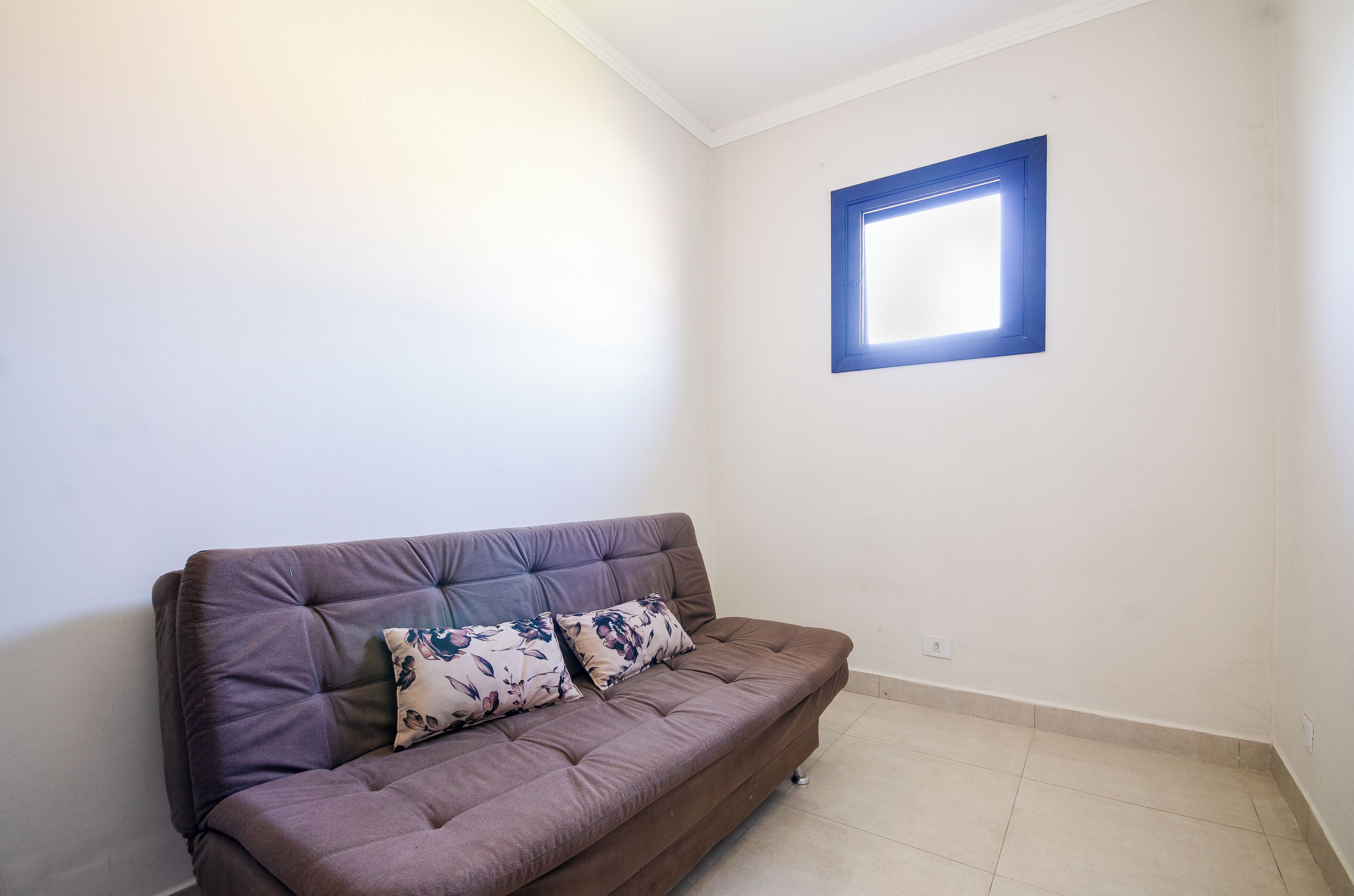 Un sofá marrón con cojines estampados en una habitación con paredes blancas y una ventana pequeña con marco azul.