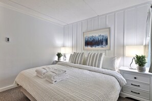 Ein ordentlich bezogenes Bett mit einem gefalteten Handtuch, ein Nachttisch mit einer Lampe, ein gerahmtes Bild und ein Blumentopf.