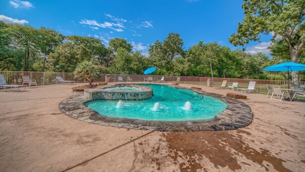 Une piscine avec un jacuzzi, entourée de chaises longues et de parasols, le tout dans un cadre verdoyant avec des arbres en arrière-plan et un ciel dégagé.
