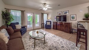 Un salon spacieux avec un ventilateur de plafond, une table basse en verre, un canapé, un téléviseur et un meuble.
