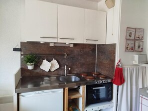 A compact kitchen with a stove, sink, and oven. There are cabinets above the counter, and a small shelf with a plant and a cloth.