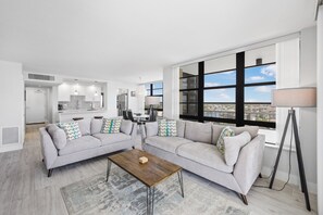 A modern living room with a grey sofa set, a wooden coffee table, and a view of a cityscape through large windows.