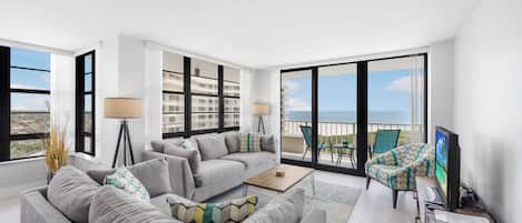 A modern living room with a grey sofa, a coffee table, and a TV. There are large windows offering a view of a building and the sky.