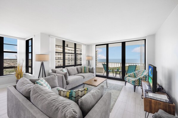 A modern living room with a grey sofa, a coffee table, and a TV. There are large windows offering a view of a building and the sky.