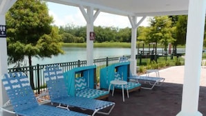 A covered outdoor seating area with blue chairs and tables, a fenced lake view, and a sign indicating a restroom. - Villas at Seven Dwarfs 4BR, 3B, WiFi, 15min DW (Kissimmee)