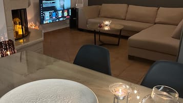Una sala de estar moderna con un sofá color beige, una mesa de centro de cristal y una mesa de comedor con un plato decorativo. Un televisor de pantalla plana está montado en la pared, y hay una chimenea con una vela encendida sobre la mesa.
