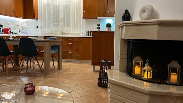 A modern kitchen with wooden cabinets, a dining area with a glass table, and a fireplace with lanterns. - Olive Gem 2-bed Apt Sleeps 4 With Fireplace (Trifylia)