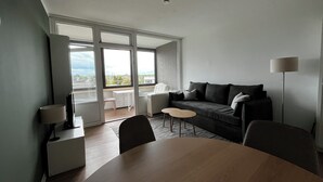 A modern living room with a large window, a sofa, a dining table, and a lamp. - Charming apartment by the sea! (Wendtorf)