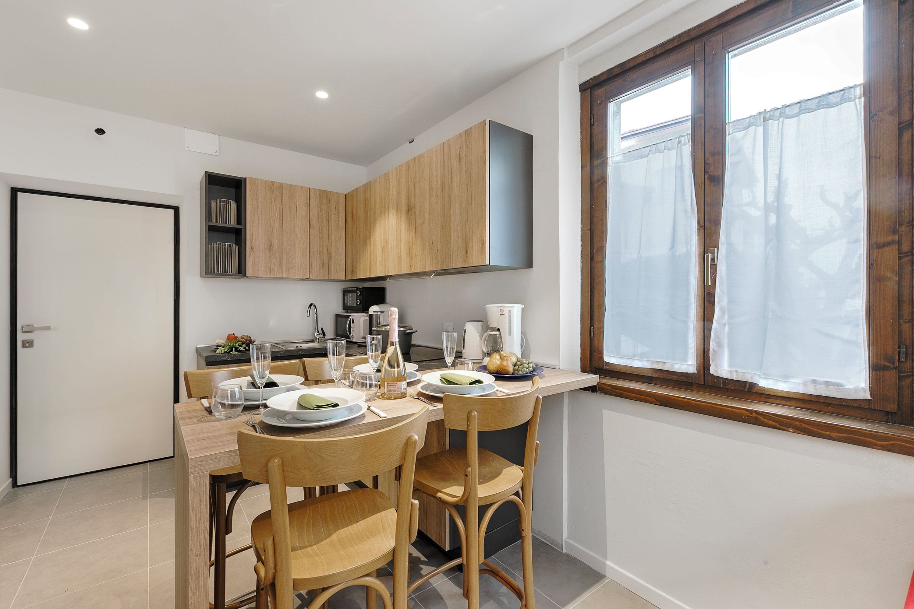 A modern kitchen with wooden cabinets, a dining table set for four, and a window with sheer curtains.