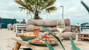 En matplats vid stranden med ett bord, tallrikar med mat, ett glas vitt vin och en soffa.