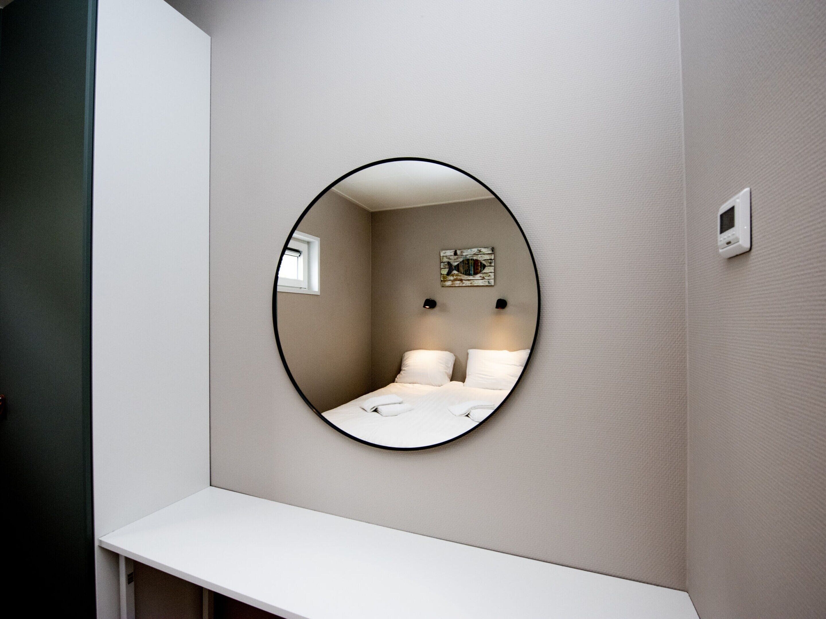 A circular mirror on a wall with a bed and a small window visible in the reflection.
