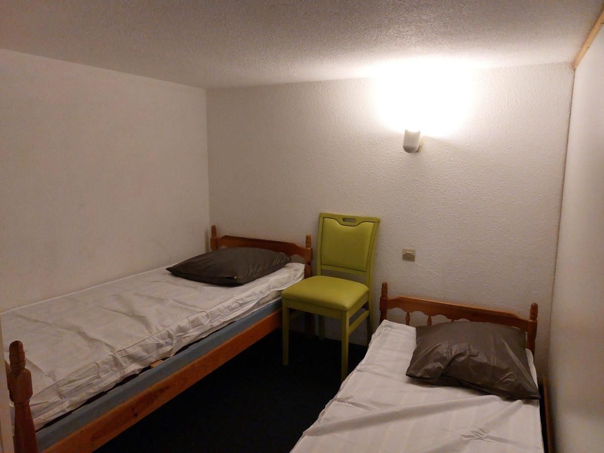A small, single-room accommodation with two single beds, a chair, and a wall-mounted light fixture.