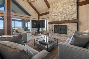 A modern living room with a stone fireplace, large windows, and a flat-screen TV.
